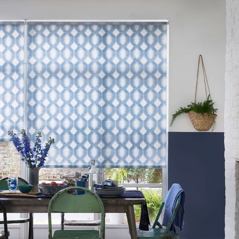 Blue patterned roller blind lowered, geometric oval motif, softly filtering daylight; above rustic dining table with mismatched chairs, vase of blue flowers, stacked dishes, hanging wicker plant, small garden visible.