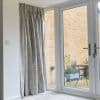 Floor-length pleated grey curtains pulled closed over the left sidelight of glass patio doors, fabric forming folds; bright daytime living room overlooking small brick patio with potted plants and table.