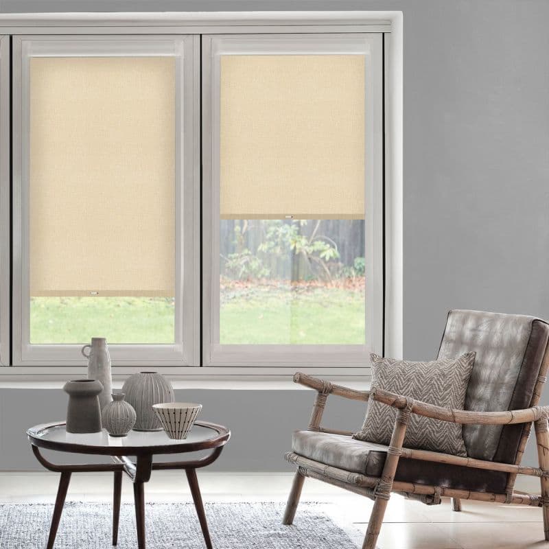 Two beige roller blinds (inside-mounted textured fabric with small pull tabs) — left fully lowered, right halfway — filtering daylight in a grey-walled living room with rattan armchair and coffee table.