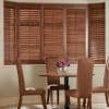 Wooden horizontal blinds (bay window) closed, slats slightly filtering light; a small dining area with round table, four brown upholstered chairs and glass vase of purple flowers against neutral walls.