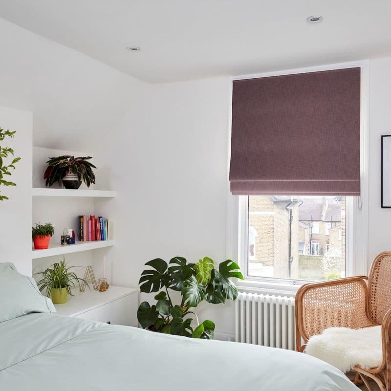 Mauve Roman blind, lowered over a tall window with fold detail, filtering daylight; in bright white bedroom with built-in shelves, plants, rattan chair and radiator, views of nearby brick houses.