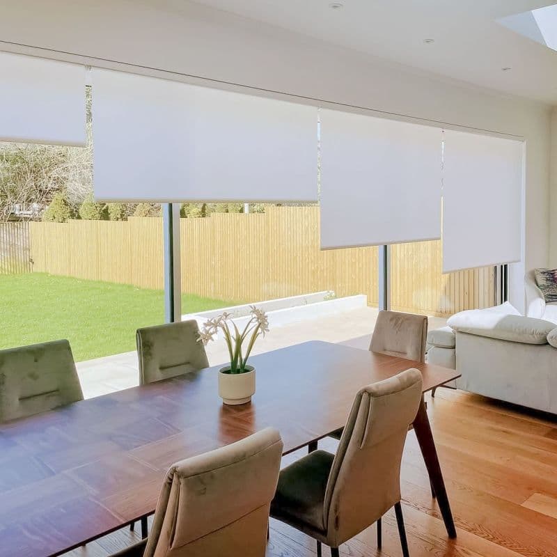 White roller blinds, four adjacent panels lowered at staggered heights, partially filtering daylight over floor‑to‑ceiling windows; bright open dining-living area with wooden table, upholstered chairs, potted plant and lawn view.
