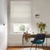 A beige fabric Roman blind, lowered with soft folds, filtering daylight into a home desk area with a wooden table, teal chair, mirror, vase of flowers, and neutral-painted walls.
