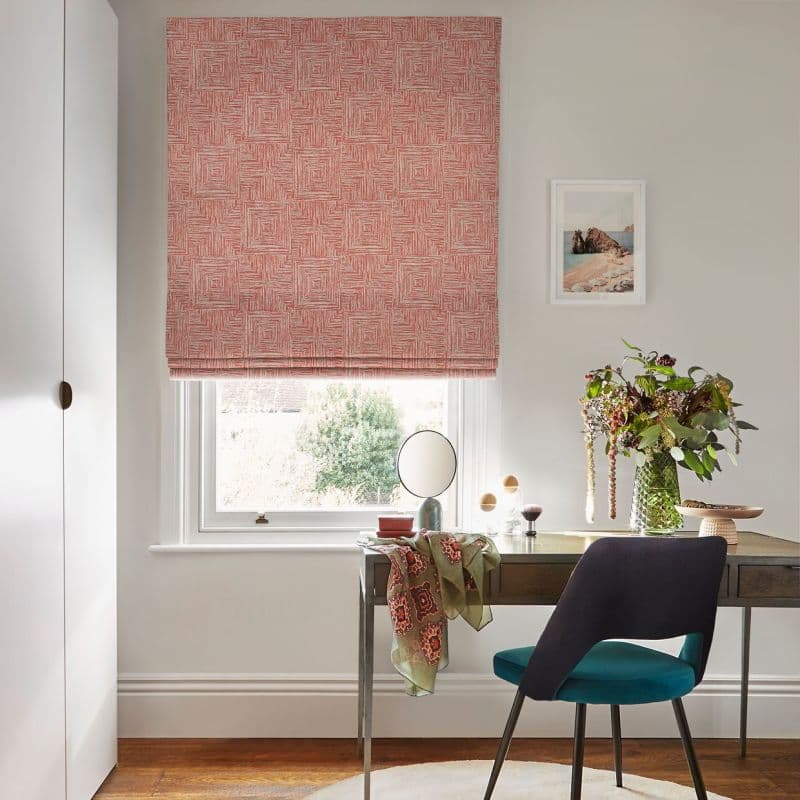Coral geometric Roman blind, lowered over a sash window, patterned with concentric square lines and filtering daylight; a small home office beneath it—wooden desk, teal chair, vase of flowers.