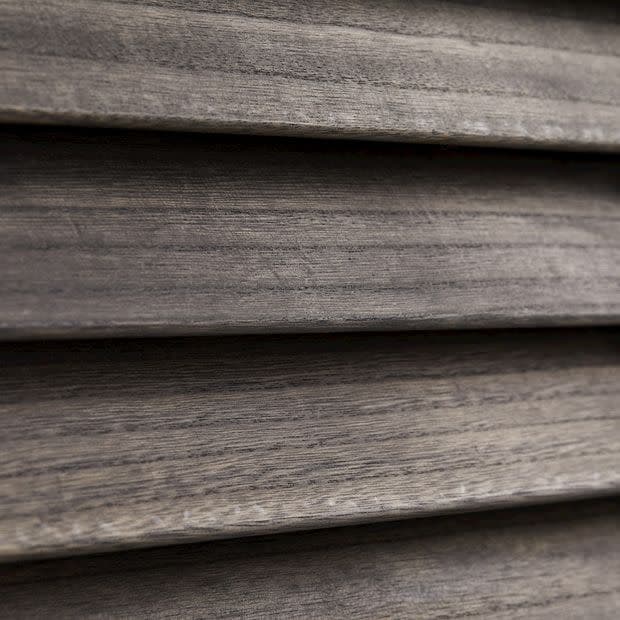 Horizontal wooden blinds, narrow slatted wood shown in close-up; closed and overlapping, textured grain visible; filtering/diffusing light in a dim interior setting with neutral gray‑brown tones.