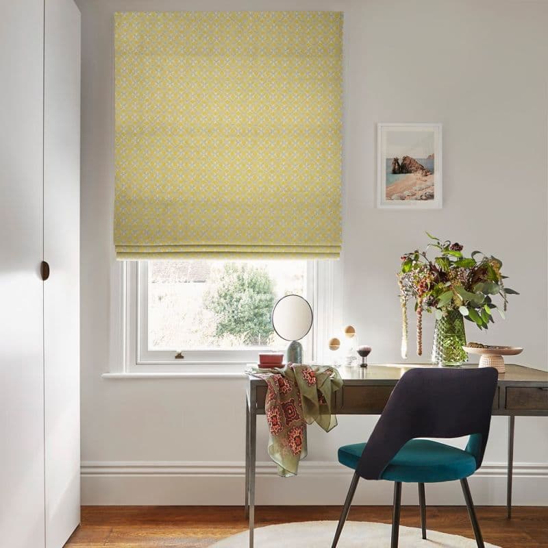 Yellow patterned Roman blind lowered over the window, softly filtering daylight; a desk with mirror, vase of flowers and teal chair fills a bright, minimalist home office overlooking a garden.