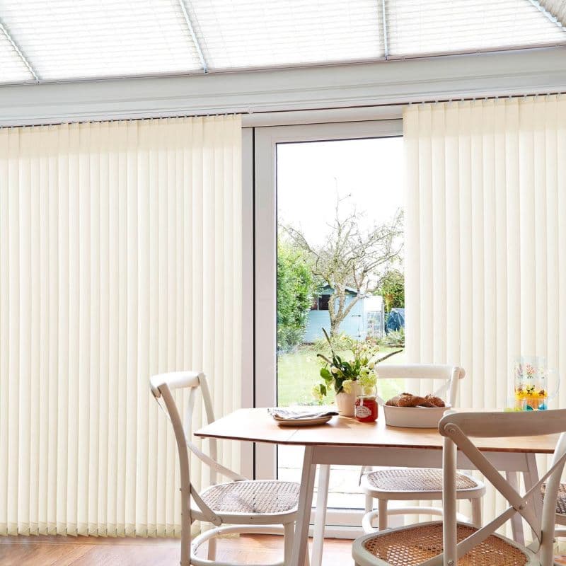 cream-colored vertical blinds cover sliding glass doors, parted at center, filtering bright daylight; small wooden dining table with white chairs, vase and bowl in sunlit dining area overlooking a garden