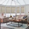Beige pleated blinds across tall bay conservatory windows, lowered and filtering soft daylight; sunlit living area with neutral sofa draped in chunky pink throw, coffee table, chest and blue rug.