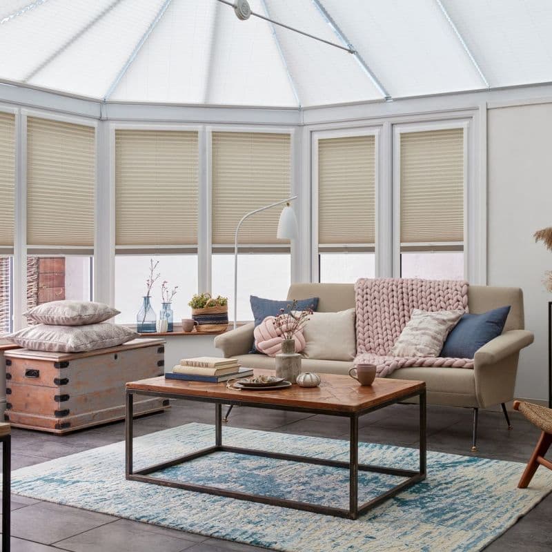 Beige pleated blinds across tall bay conservatory windows, lowered and filtering soft daylight; sunlit living area with neutral sofa draped in chunky pink throw, coffee table, chest and blue rug.