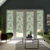 Patterned floral roller blinds cover French doors and sidelights, filtering daylight in a modern living room with green velvet chairs, glass coffee table atop black-and-white chevron rug, garden visible outside.