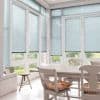 Light-blue horizontal Venetian blinds cover tall French doors and surrounding windows, lowered and filtering soft daylight; bright white dining nook with wooden table, chairs, floral cushions, tiled floor, garden beyond.
