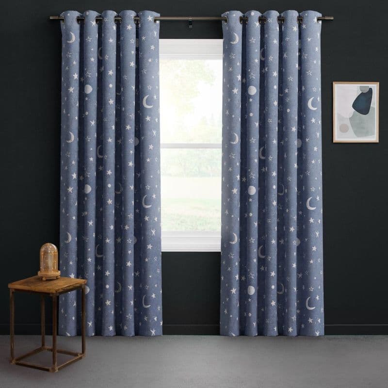 Grommet-top navy curtains patterned with moons and stars, hung on a metal rod, parted to admit soft daylight; dark-painted room with wooden side table, dome lamp, abstract print.