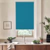 A teal roller blind covers a window, partially lowered above stainless faucet; modern kitchen sink and wooden cabinets sit below, countertop holds bowl of greens, potted plants, framed sketch.