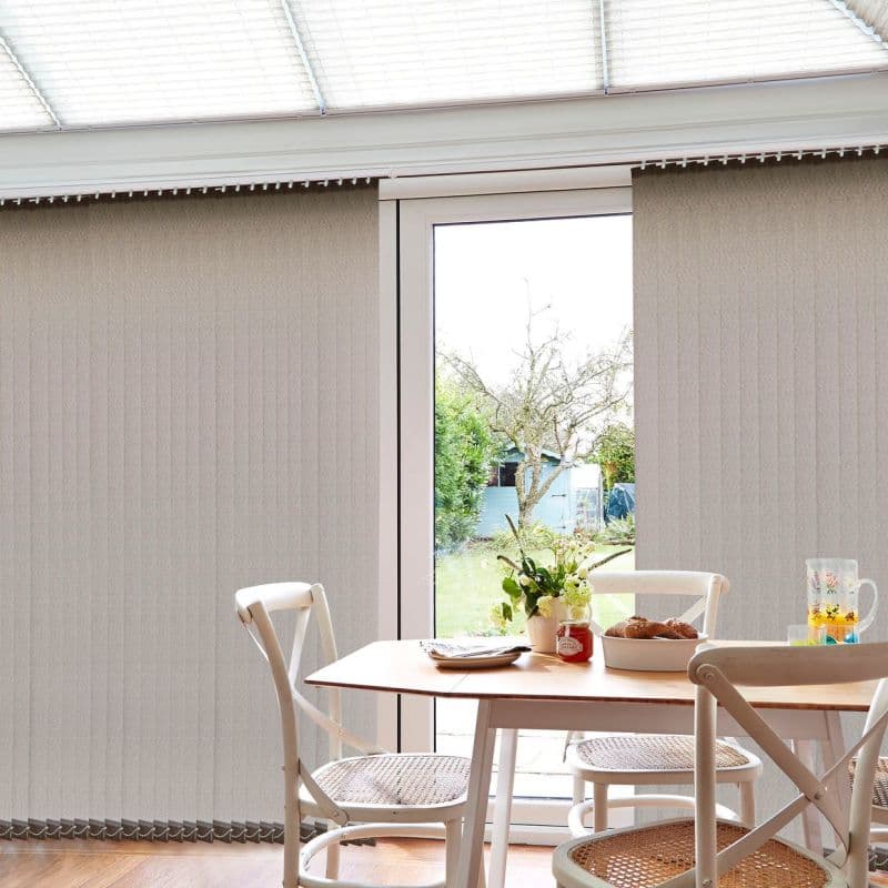 Beige vertical blinds (textured, floor‑length) cover a sliding glass door, partially opened at the center to reveal a garden; dining table with chairs sits inside a bright conservatory‑like room.