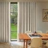 Beige textured floor-to-ceiling curtains, hung across a tall three-pane window and slightly parted, filtering soft daylight into a dining room with a wooden table, chairs, ceramic pitcher, and garden view.