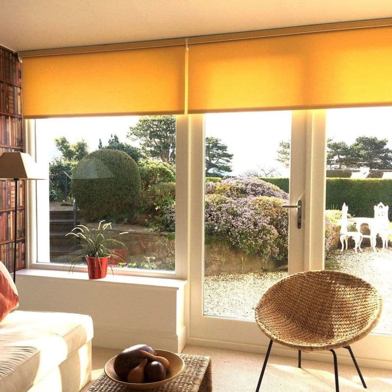 Yellow roller blinds across a wide glass door and window, lowered halfway and filtering warm sunlight; living room wicker chair, potted plant on sill, sunlit garden of trimmed shrubs outside.