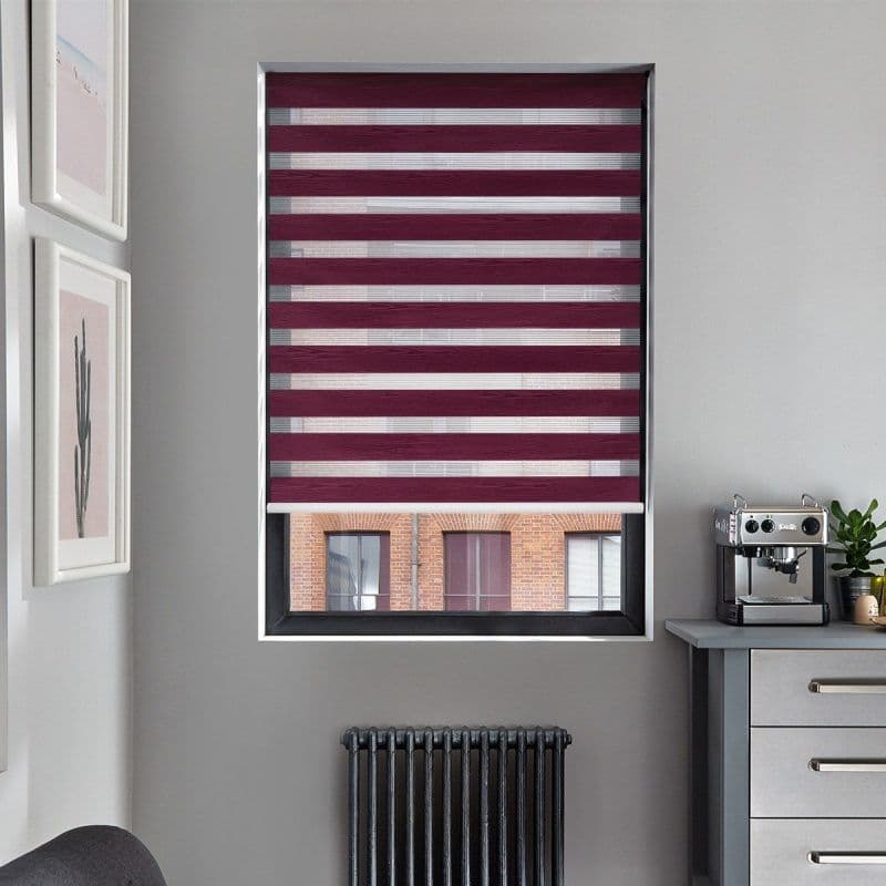 Dual-layer zebra roller blind (burgundy opaque and sheer horizontal stripes) fully lowered across window, filtering light and revealing partial view of a brick building; gray kitchen corner with radiator.