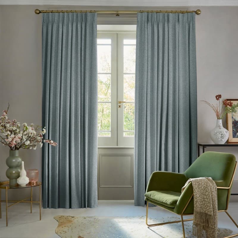 Pleated blue-gray curtains on a brass rod, hung full-length and parted at center, filtering daylight and framing a paneled French window in a softly lit living room with green armchair.