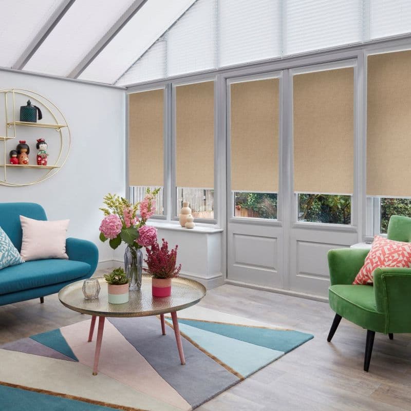 Beige roller blinds lowered mid-height across tall conservatory windows and doors, partially blocking and diffusing daylight, in a bright sunroom with teal sofa, green armchair, round coffee table and flowers.