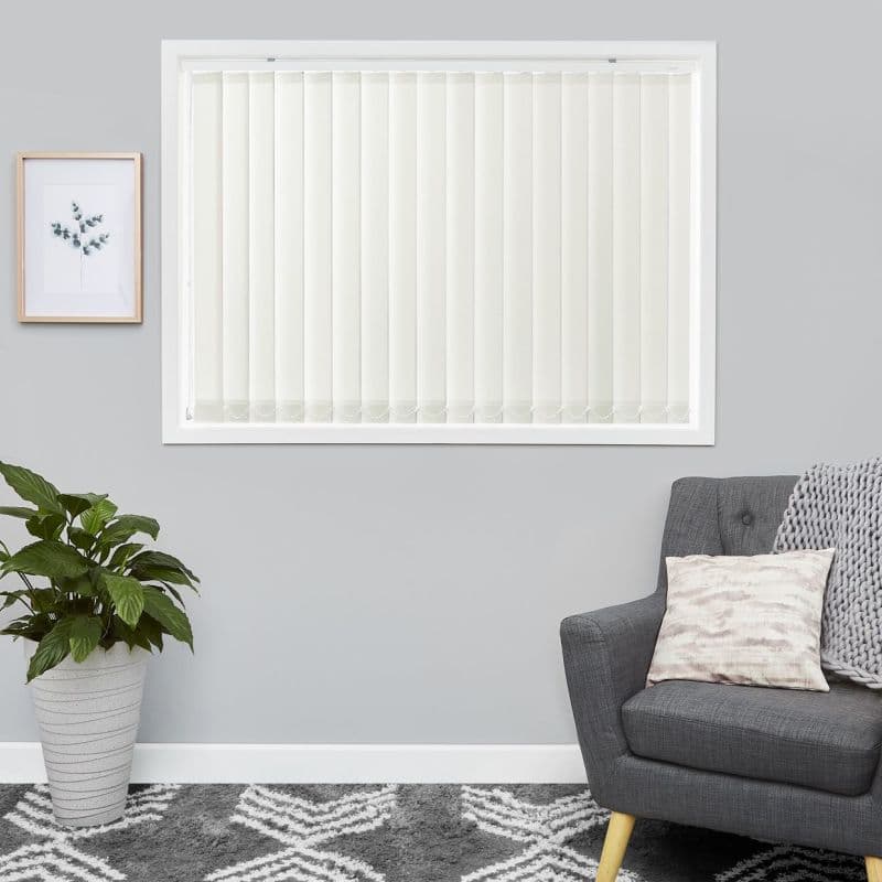 Vertical off-white slatted blinds covering a window, closed and softly filtering daylight; in a living room with grey walls, a grey armchair with cushion and throw, potted plant, framed print.