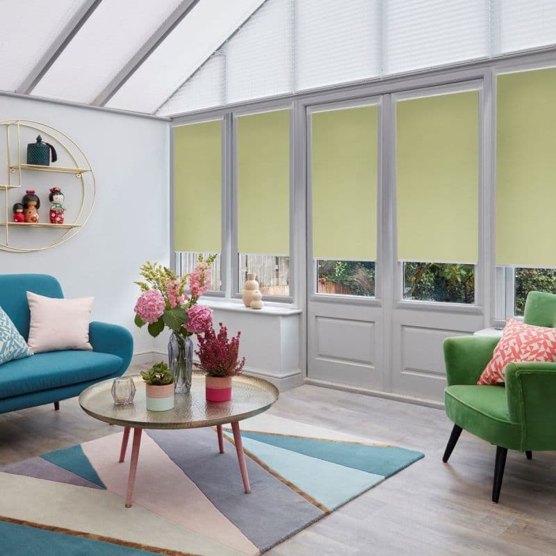 Pale green roller blinds lowered over tall conservatory windows and glass doors, filtering light; sunroom with teal and green armchairs, geometric pastel rug, round coffee table and vases of flowers.