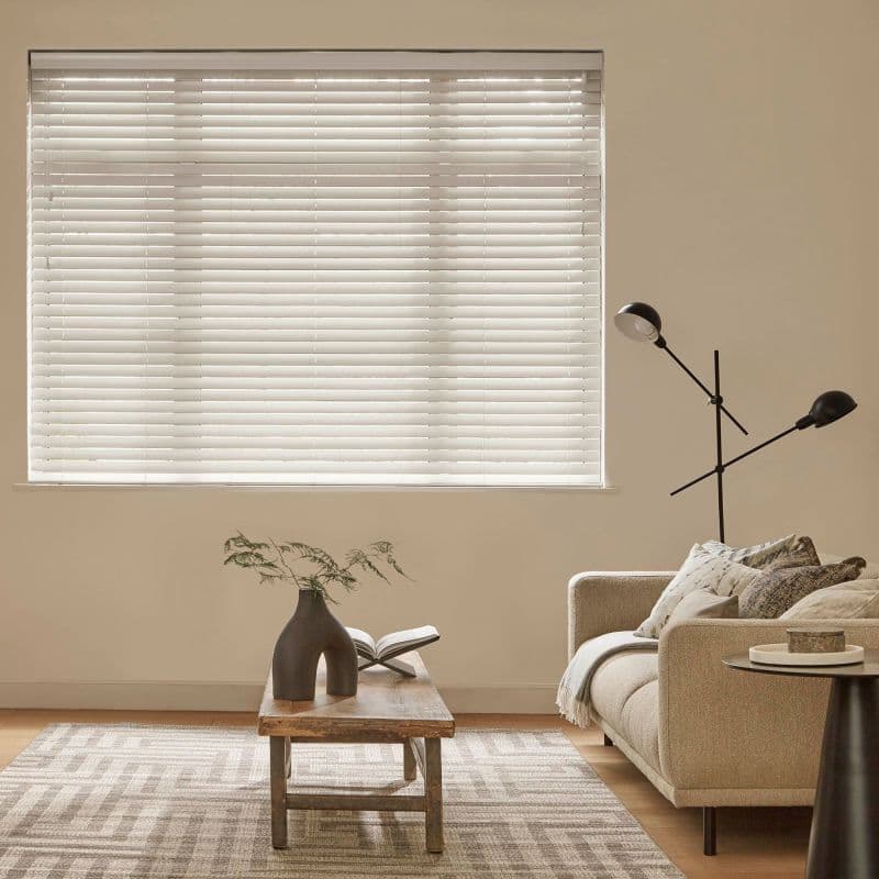 White horizontal blinds covering a large rectangular window, fully lowered with slats closed, softly filtering daylight; in a neutral living room with beige walls, sofa, wooden coffee table and lamp.