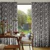 Black-and-white botanical print full-length curtains, pleated and pulled aside to frame a sliding glass door; filtering soft morning light onto a dining table set for breakfast, with a garden visible beyond.