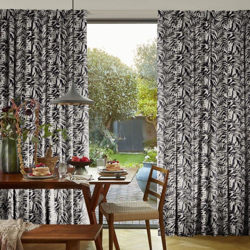 Black-and-white botanical print full-length curtains, pleated and pulled aside to frame a sliding glass door; filtering soft morning light onto a dining table set for breakfast, with a garden visible beyond.