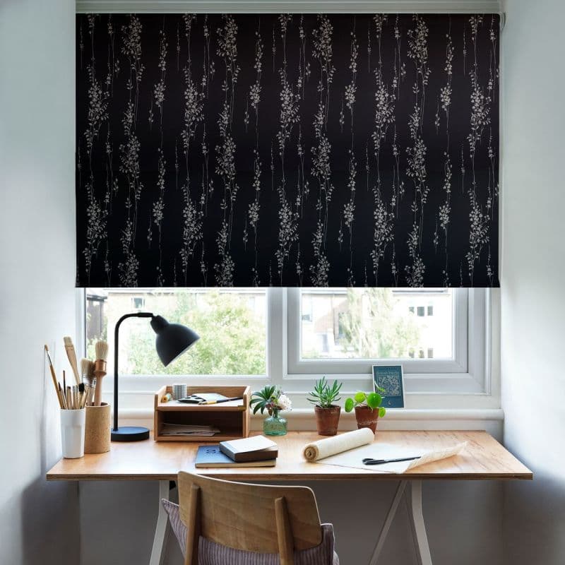 Black roller blind with pale vertical floral print, lowered over a double window, filtering light; wooden desk beneath holds a lamp, paintbrushes, notebooks and potted succulents, view of nearby houses.
