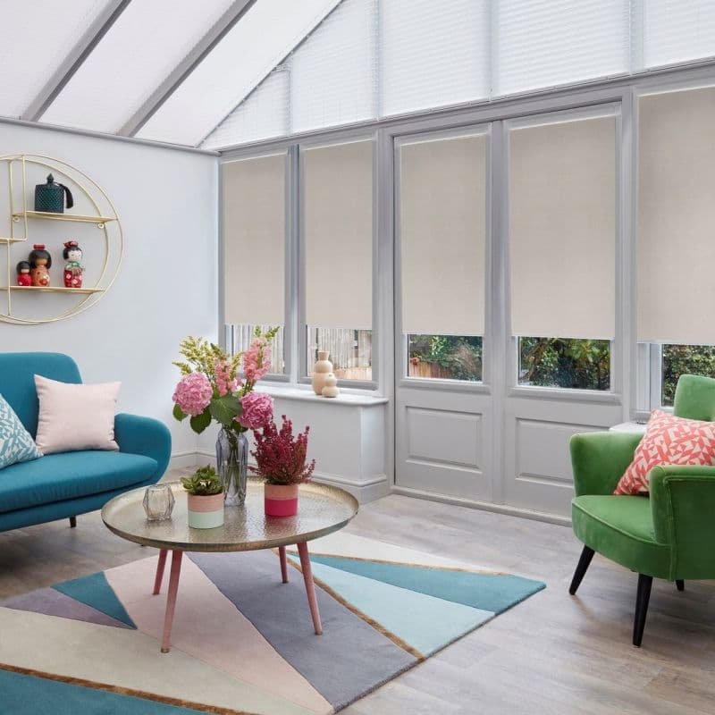Beige roller blinds covering tall glass doors and windows, lowered halfway and filtering daylight; conservatory roof has pleated blinds—bright sunroom with colorful sofas, a coffee table, and potted flowers.