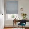Grey Roman blind, lowered and partially filtering daylight over a sash window; a desk beneath holds mirror, vase of flowers and draped scarf, with teal chair in a study.