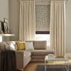 Cream pleated curtains flank a patterned leaf roller blind, the blind partially lowered and filtering daylight; in a neutral living room with a beige sectional, yellow and white cushions, coffee table, shelf.