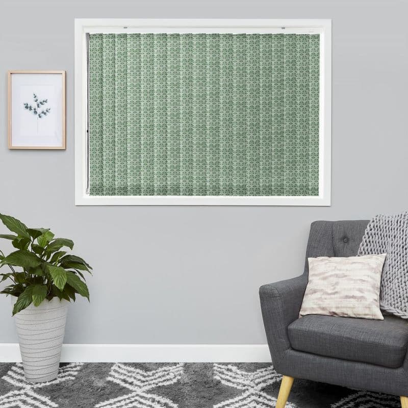 Green geometric-patterned pleated blind, fully closed and filtering light; in a grey-walled living room with framed leaf print, potted plant, grey armchair, knitted throw and patterned rug.