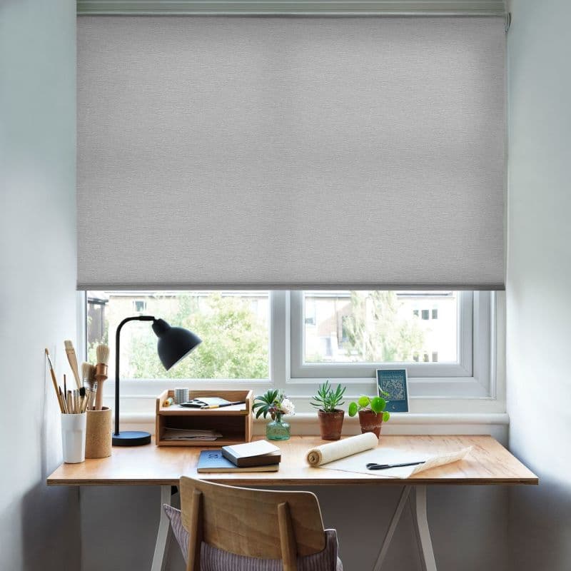 Light-gray fabric roller blind, fully lowered across the window, smooth texture; filtering soft daylight into a small home desk area with lamp, brushes, notebooks and three potted plants.