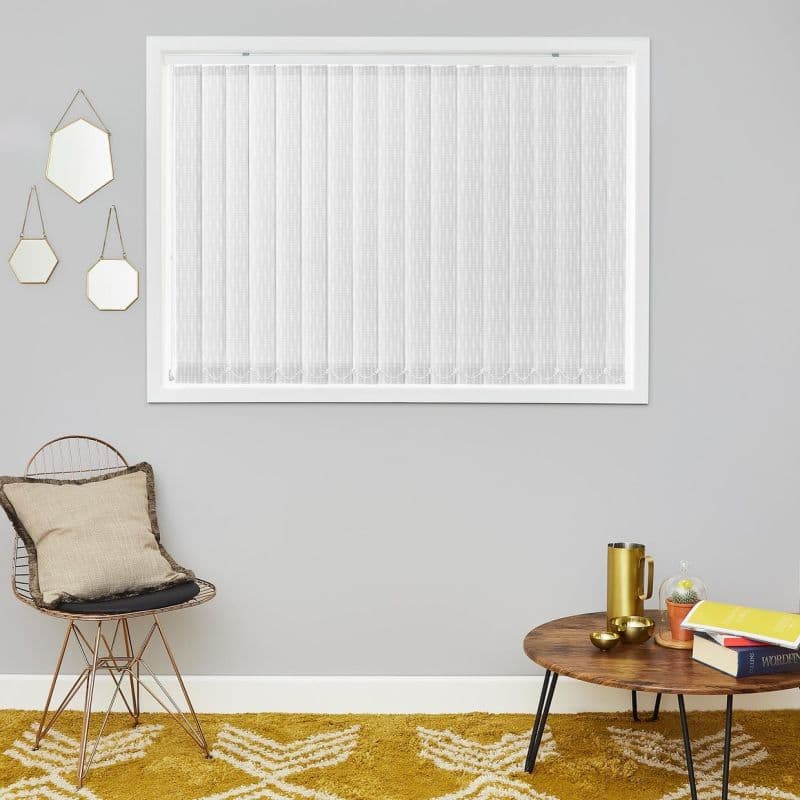 White vertical blinds (full-window, narrow textured slats) — closed, diffusing daylight — set in a grey-walled living space with hanging geometric mirrors, wire chair with cushion, wooden coffee table, mustard rug.