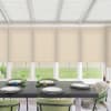 Dining table set with plates, small colored candles and green glass vases sits in a bright sunroom with beige roller blinds partly lowered, green chairs, and a grassy yard outside.