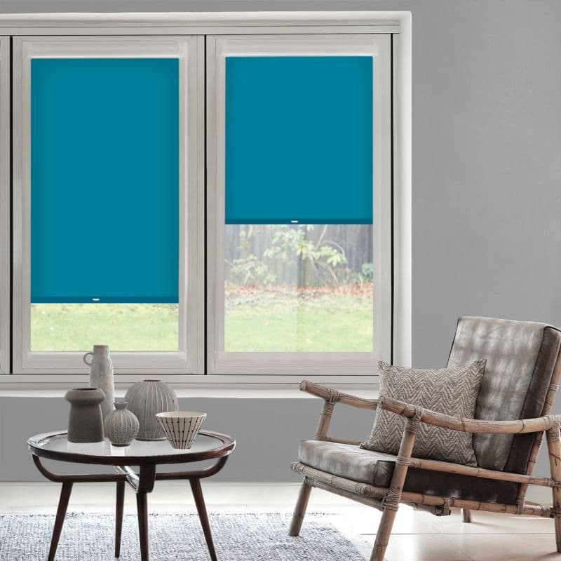 Two teal roller blinds on adjacent windows, smooth fabric—left fully lowered, right half-lowered—partially blocking light and revealing a lawn; gray living room with bamboo-framed leather chair and coffee table.