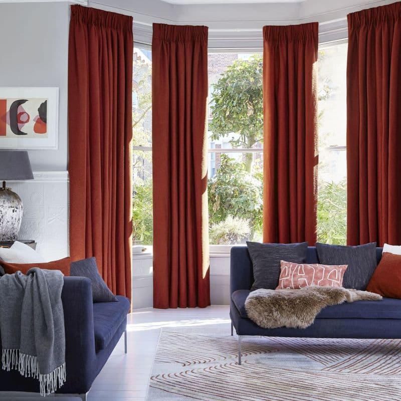 Rust-red pleated floor-length curtains across a bay window, partially closed and filtering sunlight; modern living room with navy sofas, patterned rug, lamp, and a leafy garden visible outside.