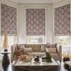 Patterned Roman shades (mauve-gray diamonds) cover three bay windows, lowered to softly filter light and partially obscure the view; neutral living room with tufted beige sofa, marble coffee table, plants.