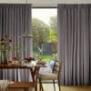 Grey pleated floor-to-ceiling curtains, slightly parted at center revealing glass door and garden; filtering soft daylight. Dining table set with pancakes and a pendant lamp in a bright dining area.