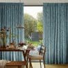 Teal textured, floor-length pleated curtains pulled apart to frame a sunlit garden view, filtering light; a wooden dining table set with pancakes, vases and a hanging pendant lamp.