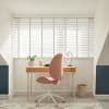 White horizontal slat blinds with vertical tapes covering the full window, closed and softly diffusing bright daylight, framing a small, sunlit home office alcove with wooden desk, pink swivel chair.