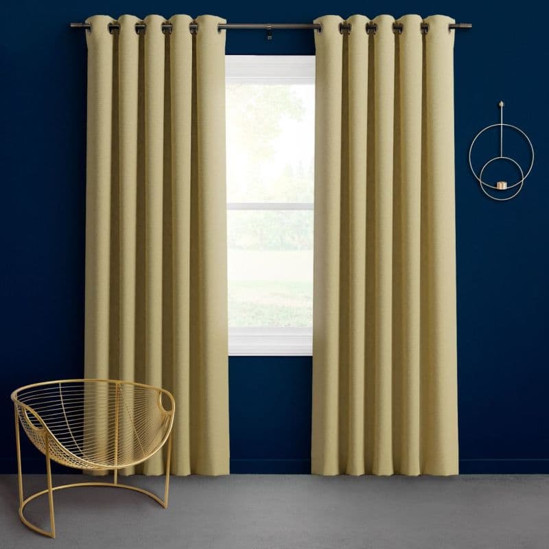 Grommet-top beige curtains hung on a metal rod, pulled aside to reveal a bright window in neat pleats; against a deep navy wall with a gold wire chair and circular wall decor.