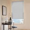 Light-gray fabric roller blind, fully lowered with a small bottom gap, filtering soft daylight over a minimalist home-office/vanity corner with pale walls, black curved desk, and a wire chair.