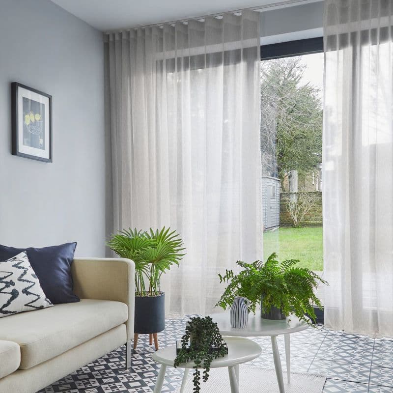 Sheer beige, ceiling-mounted curtains—full-height with soft vertical folds—slightly parted, filtering daylight; in a bright living room beside a pale sofa and patterned tile floor, potted plants foreground, garden visible.