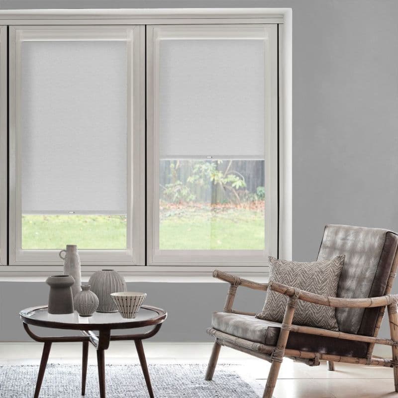 White fabric roller blinds, inside-mounted over a two-panel window—left fully lowered, right half-lowered—filtering soft daylight. Neutral living room with gray walls, rattan-framed leather armchair, round table and garden view.