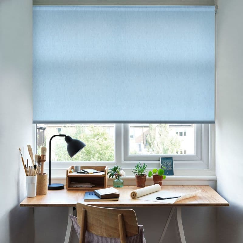 Light-blue roller blind, textured fabric, lowered over a double window; filtering soft daylight. A wooden desk beneath holds a lamp, brushes, notebooks, and small potted succulents.