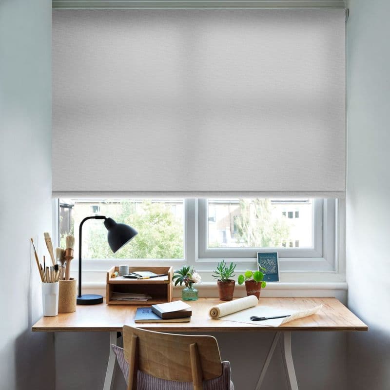 Light-gray roller blind lowered across the window, diffusing daylight. Below, a wooden desk with a lamp, notebooks, art brushes, small potted plants and workspace items in a bright home study.