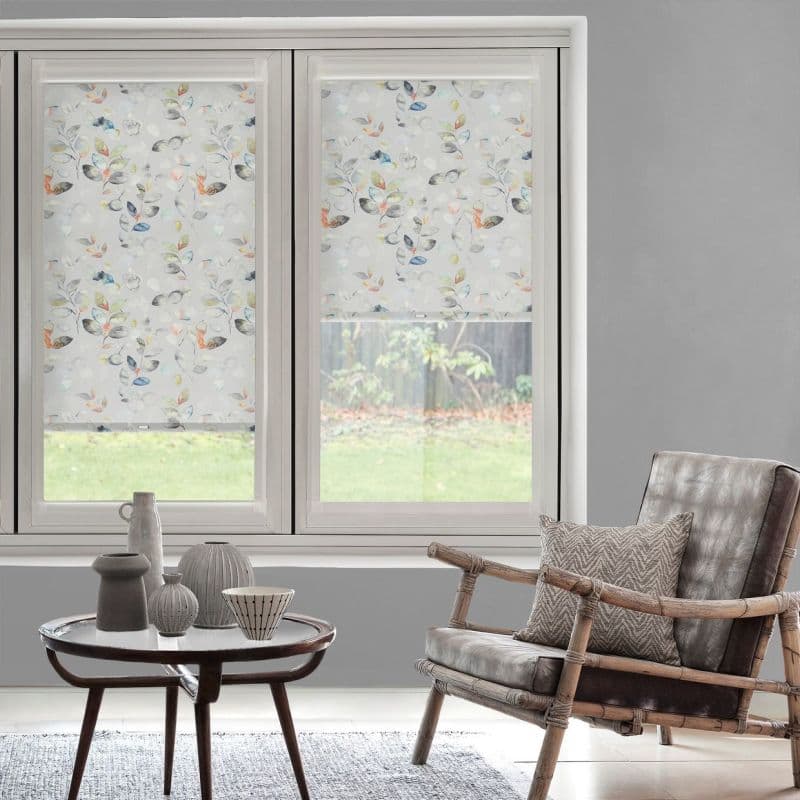 Two translucent roller blinds with watercolor leaf print cover a double window — left down, right partially raised, filtering soft daylight in a neutral living room with rattan armchair, round table.
