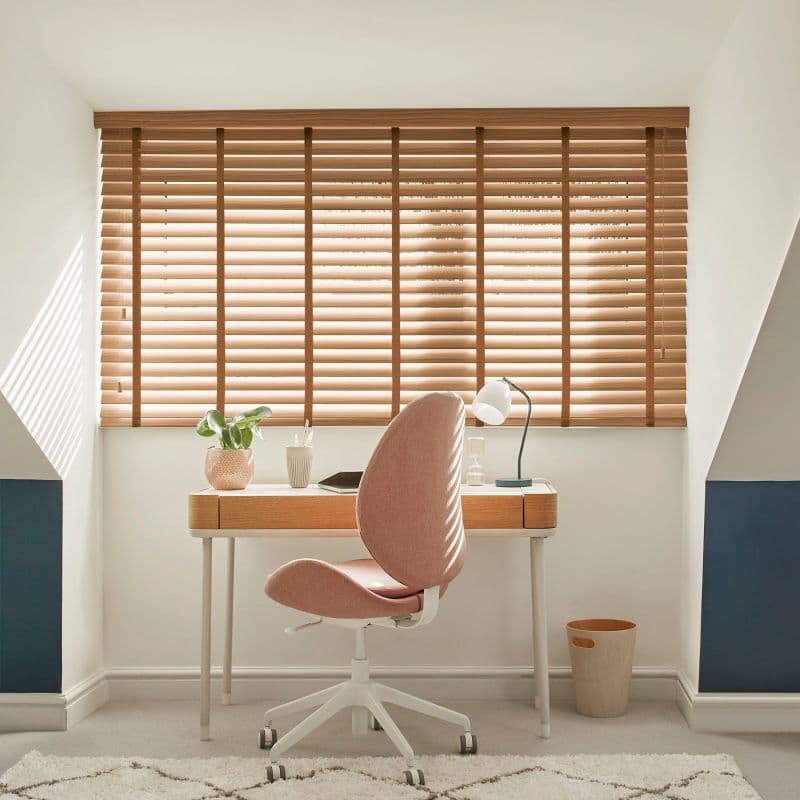 Wooden horizontal blinds covering a wide dormer window, slats tilted to filter sunlight; a compact desk with lamp, plant and pink office chair in a bright attic study.
